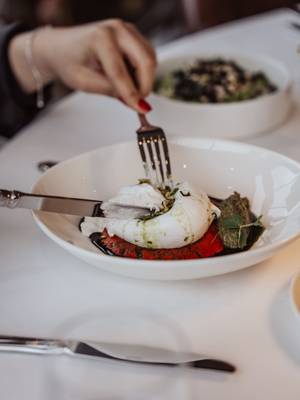 Close-up of a hand with red nail polish cutting into a ball of Burrata cheese served with balsamic glaze and pesto on a red base at Vetiver Restaurant, Rixos Pera Istanbul.