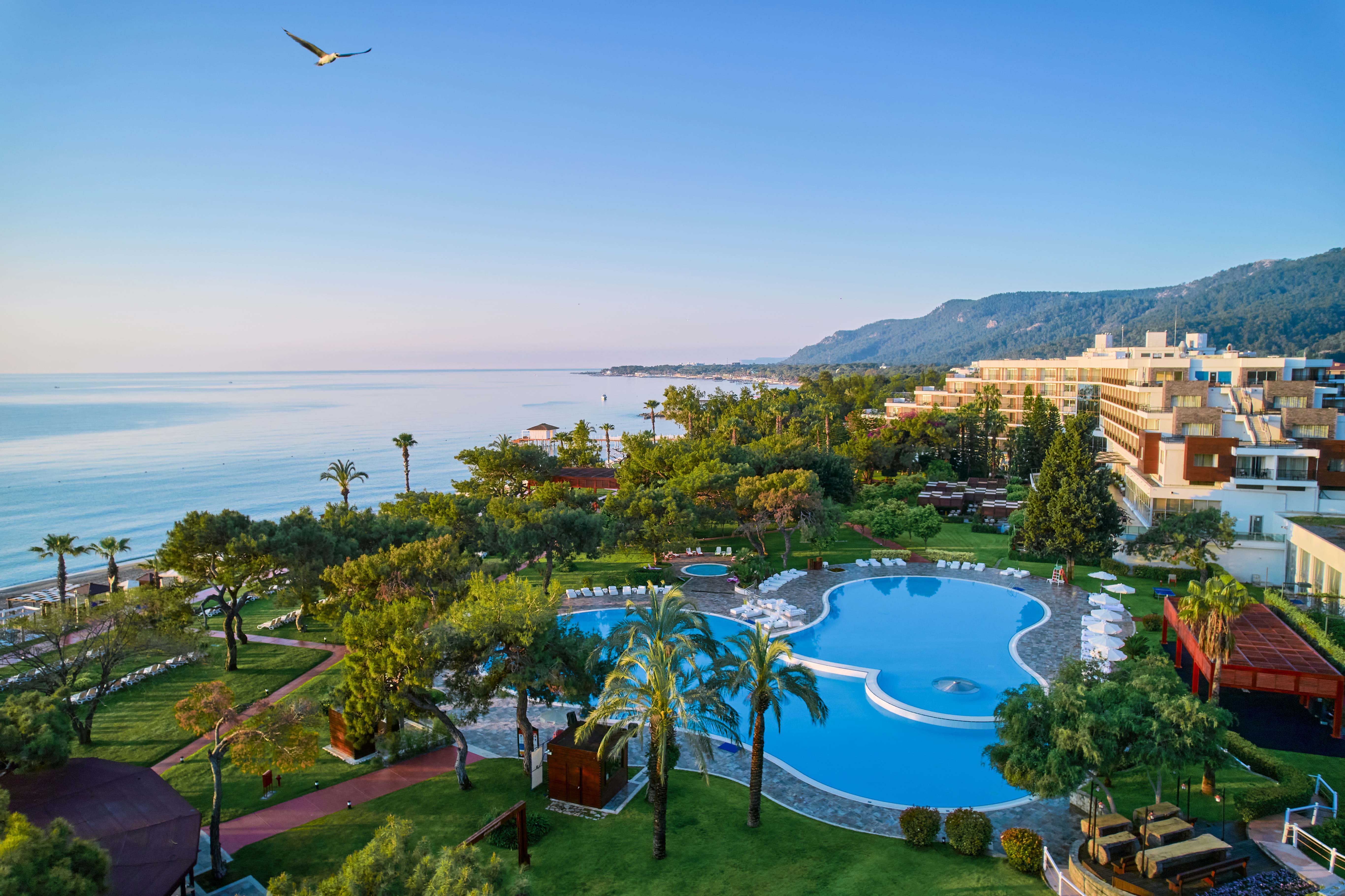 Aerial view of Rixos Beldibi, a coastal resort with a large circular swimming pool, palm trees, lounge chairs, and adjacent modern buildings, set by the sea with mountains in the background